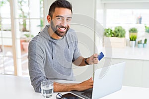 Handsome man smiling using credit card as payment metod when shopping online using laptop