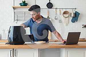 Handsome man standing at kitchen table and cooking toast bread healthy with recipe by Air Fryer machine, using laptop computer