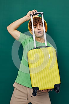 a handsome man stands on a green background, dressed in a yellow panama hat, holding a bright travel suitcase in his