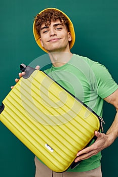 a handsome man stands on a green background, dressed in a yellow panama hat, holding a bright travel suitcase in his