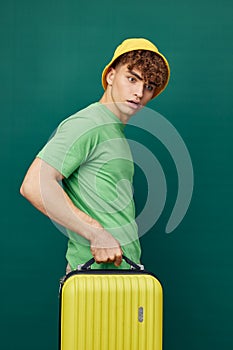 a handsome man stands on a green background, dressed in a yellow panama hat, holding a bright travel suitcase in his