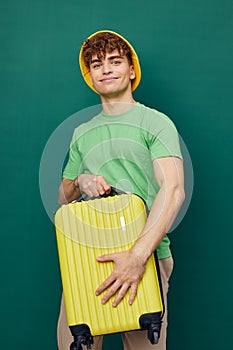 a handsome man stands on a green background, dressed in a yellow panama hat, holding a bright travel suitcase in his