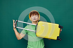 a handsome man stands on a green background, dressed in a yellow panama hat, holding a bright travel suitcase in his