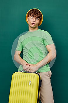 a handsome man stands on a green background, dressed in a yellow panama hat, holding a bright travel suitcase in his