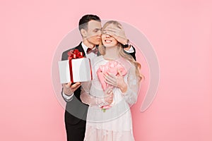 Handsome man in a suit making a surprise to a woman, closing her eyes with her hands on a pink background, concept of women`s day