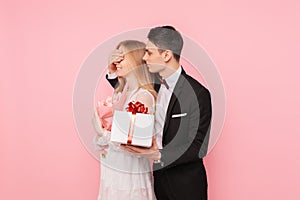 Handsome man in a suit making a surprise to a woman, closing her eyes with her hands on a pink background, concept of women`s day