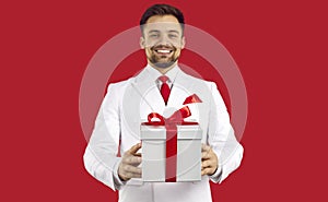 Handsome man in a suit standing on a red studio background holding a white present box