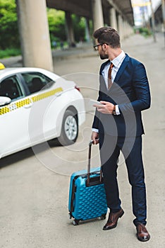 handsome man with suitcase and smartphone looking at taxi cab
