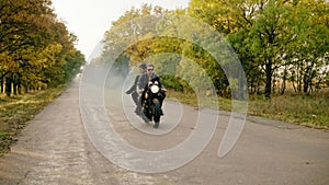 Handsome man in sunglasses riding with his girlfriend on a motorcycle on the asphalt road in forest in autumn. His