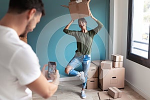 Handsome man taking a photo with smart phone to his funny wife while she jokes with cardboard boxes in the new house