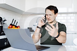 Handsome man talking on mobile phone while using laptop at the table at home