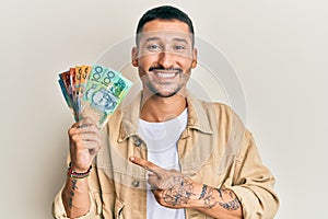 Handsome man with tattoos holding australian dollars smiling happy pointing with hand and finger