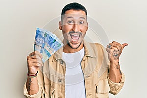 Handsome man with tattoos holding south african 100 rand banknotes pointing thumb up to the side smiling happy with open mouth