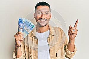 Handsome man with tattoos holding south african 100 rand banknotes smiling happy pointing with hand and finger to the side