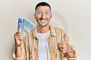 Handsome man with tattoos holding south african 100 rand banknotes smiling happy and positive, thumb up doing excellent and