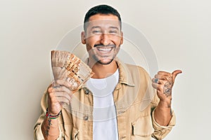 Handsome man with tattoos holding south african 20 rand banknotes pointing thumb up to the side smiling happy with open mouth
