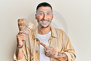 Handsome man with tattoos holding south african 20 rand banknotes smiling happy pointing with hand and finger