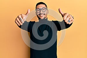 Handsome man with tattoos wearing turtleneck sweater and glasses approving doing positive gesture with hand, thumbs up smiling and