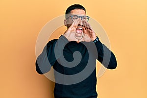 Handsome man with tattoos wearing turtleneck sweater and glasses shouting angry out loud with hands over mouth