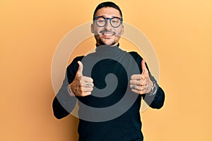 Handsome man with tattoos wearing turtleneck sweater and glasses success sign doing positive gesture with hand, thumbs up smiling