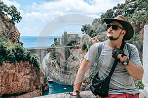 The handsome man traveler enjoys a wonderful view of the beach and the stone bridge. Sunglasses and a kangaroo leather cowboy hat