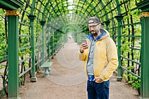 Handsome man in trendy cap, jeans and yellow anorak wearing eyeglasses holding his hand in pocket communicating online with his fr