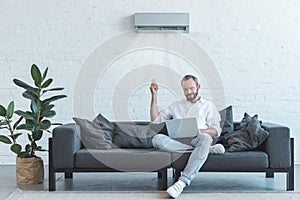 handsome man turning on air conditioner with remote control while using laptop on sofa