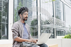 Handsome man using laptop in a city. Smiling indian male student working on computer outdoor