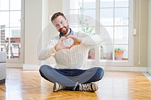 Handsome man wearing casual sweater sitting on the floor at home smiling in love showing heart symbol and shape with hands