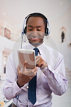 Handsome man wearing headphones with microphone, white striped shirt and tie, posing holding tablet in hand, smiling
