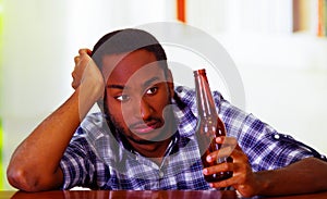 Handsome man wearing white blue shirt sitting by bar counter lying over desk holding brown beer bottle, drunk depressed