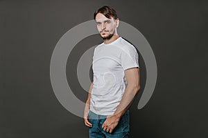 A handsome man in a white t-shirt and jeans stands on a gray background. Studio photo