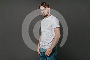 A handsome man in a white t-shirt and jeans stands on a gray background. Studio photo