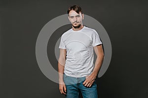 A handsome man in a white t-shirt and jeans stands on a gray background. Studio photo