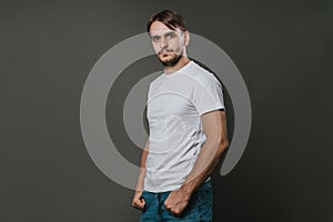 A handsome man in a white t-shirt and jeans stands on a gray background. Studio photo