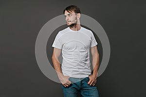 A handsome man in a white t-shirt and jeans stands on a gray background. Studio photo