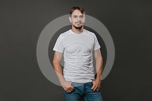 A handsome man in a white t-shirt and jeans stands on a gray background. Studio photo