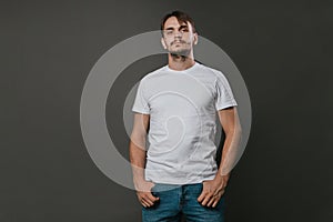 A handsome man in a white t-shirt and jeans stands on a gray background. Studio photo