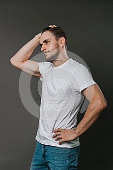 A handsome man in a white t-shirt and jeans stands on a gray background. Studio photo