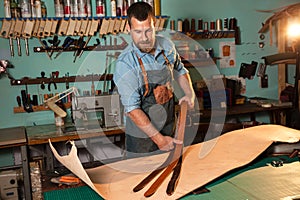 Craftsman in apron working with leather at workshop, belt manufacturing process.