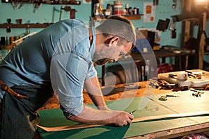 Craftsman in apron working with leather at workshop, belt manufacturing process.