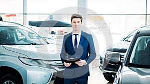 Handsome manager standing between cars in car showroom and looking