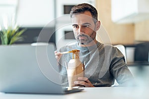 Handsome mature business man eating take away noodles while working with laptop at home