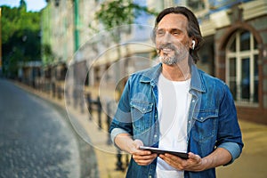 Handsome mature freelancer man with digital tablet in hands having a call while standing on urban streets. Mature man