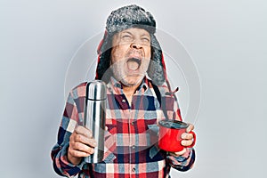 Handsome mature handyman wearing winter hat with ear flaps drinking hot coffee from thermo angry and mad screaming frustrated and