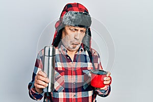 Handsome mature handyman wearing winter hat with ear flaps drinking hot coffee from thermo depressed and worry for distress,