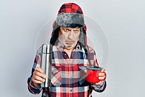 Handsome mature handyman wearing winter hat with ear flaps drinking hot coffee from thermo skeptic and nervous, frowning upset