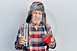 Handsome mature handyman wearing winter hat with ear flaps drinking hot coffee from thermo winking looking at the camera with sexy