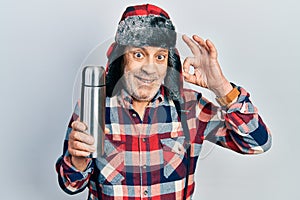 Handsome mature handyman wearing winter hat with ear flaps holding thermo doing ok sign with fingers, smiling friendly gesturing