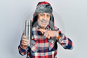 Handsome mature handyman wearing winter hat with ear flaps holding thermo smiling happy pointing with hand and finger